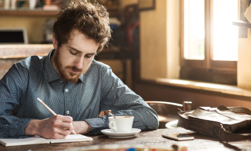 A man writing in a notebook next to a cup of coffee